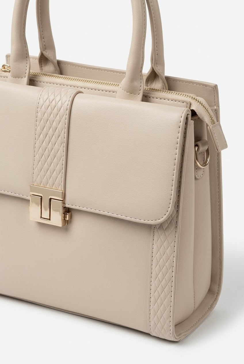 Angled close-up of cream satchel showing T-bar clasp and embossed panel